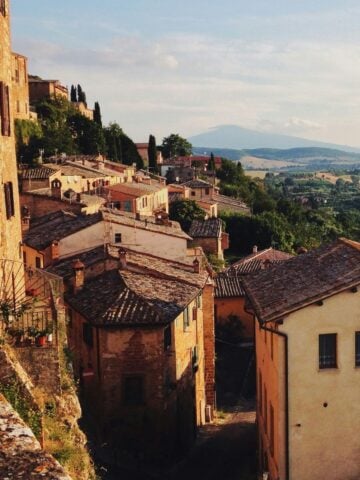 A sunlit hillside village with terracotta rooftops and rustic stone buildings, surrounded by green fields and distant mountains under a blue sky.