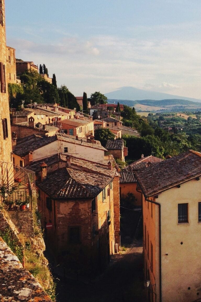 A sunlit hillside village with terracotta rooftops and rustic stone buildings, surrounded by green fields and distant mountains under a blue sky.