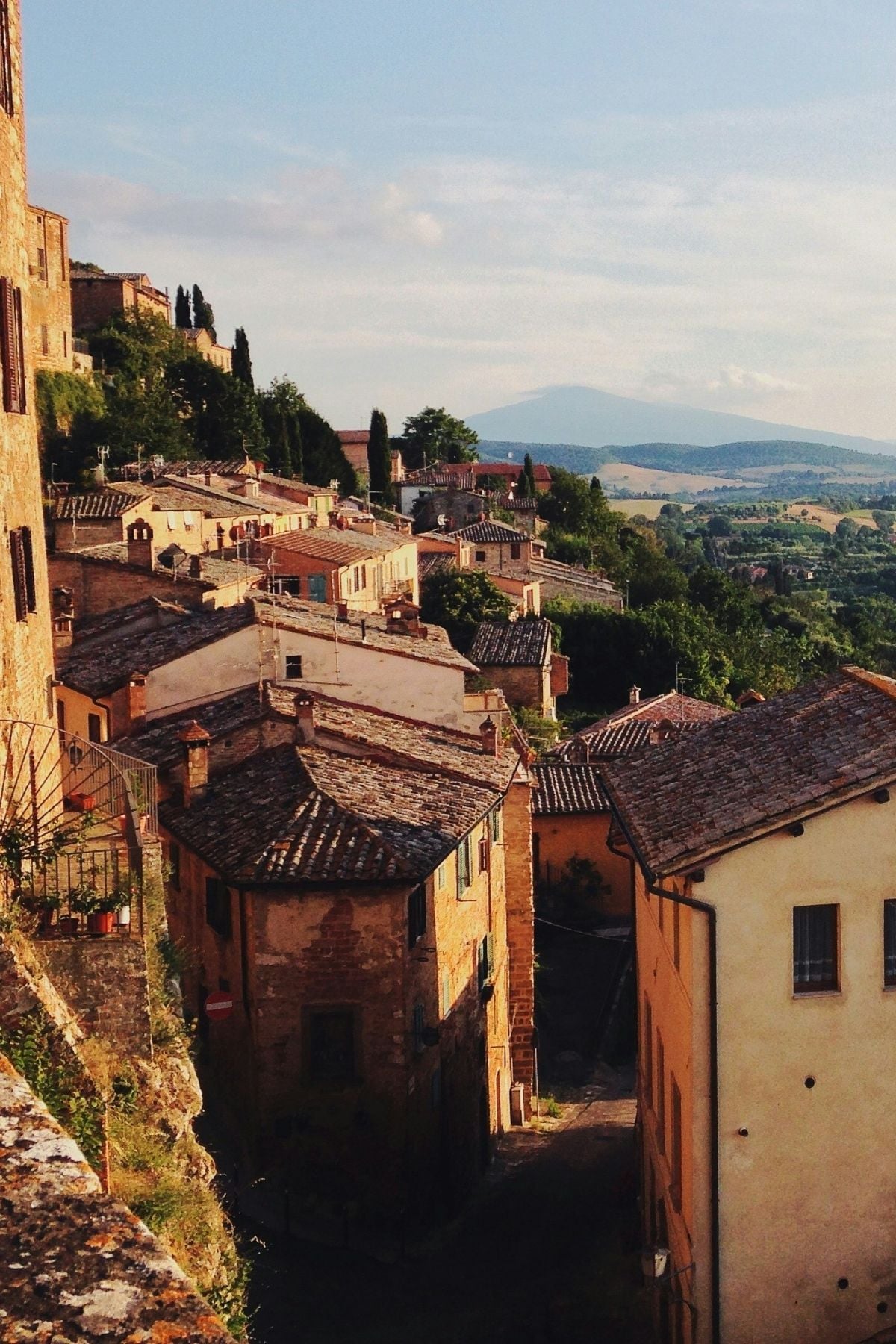 A sunlit hillside village with terracotta rooftops and rustic stone buildings, surrounded by green fields and distant mountains under a blue sky.