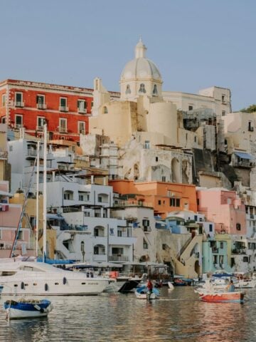 Colorful buildings and a domed church rise above a waterfront lined with boats, reflecting in the calm water under a clear sky.