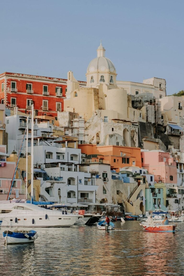 Colorful buildings and a domed church rise above a waterfront lined with boats, reflecting in the calm water under a clear sky.
