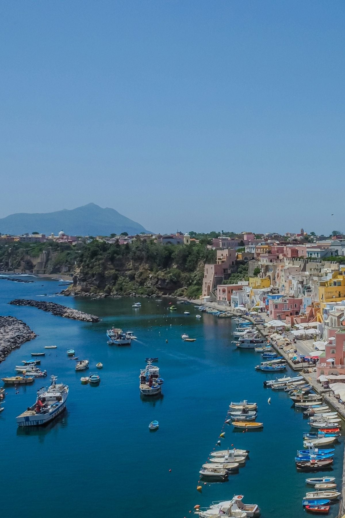 Colorful buildings line a coastal village with boats docked along a calm blue harbor. Rocky cliffs and distant mountains are visible under a clear, sunny sky.