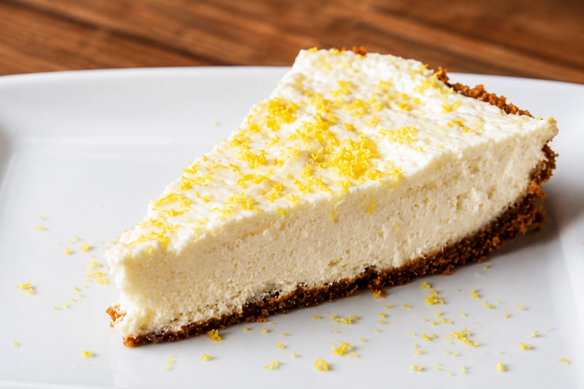 A slice of creamy No-Bake Limoncello Cheesecake with a crumbly brown crust, topped with lemon zest, served on a white plate against a wooden background.