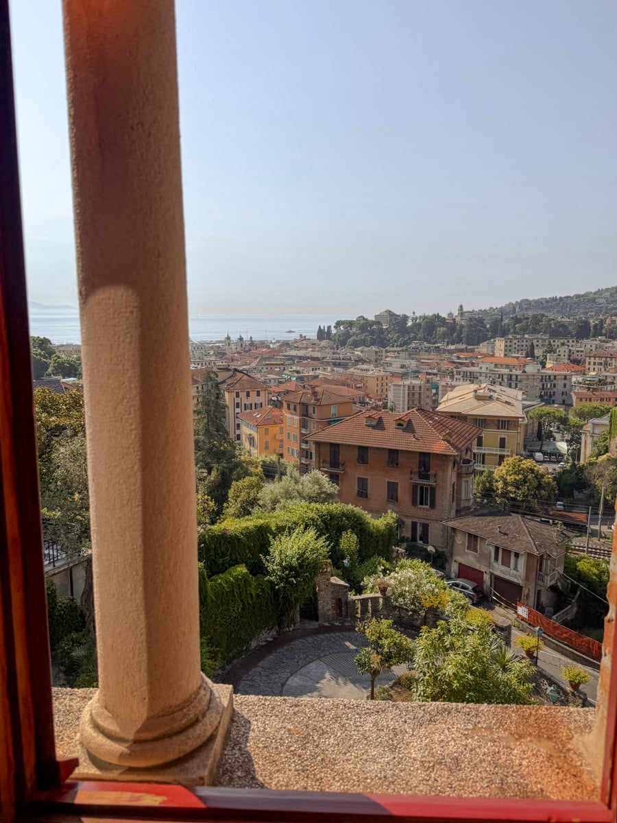 View from a window with stone columns overlooking Santa Margherita Ligure, where colorful buildings, lush trees, and the distant sea shimmer beneath a clear Mediterranean sky.
