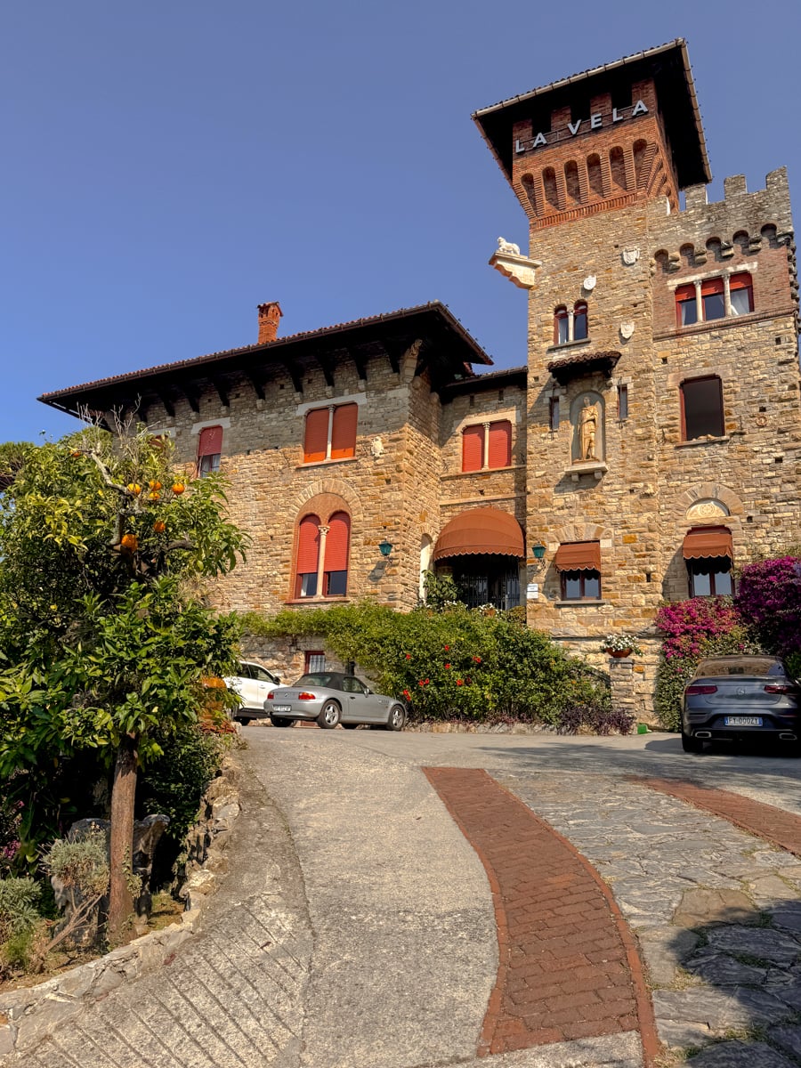 Stone castle with red-trimmed windows, a tower labeled LA VELA, lush greenery, blooming flowers, and parked cars along a curved driveway under a clear blue sky in the picturesque town of Santa Margherita Ligure.
