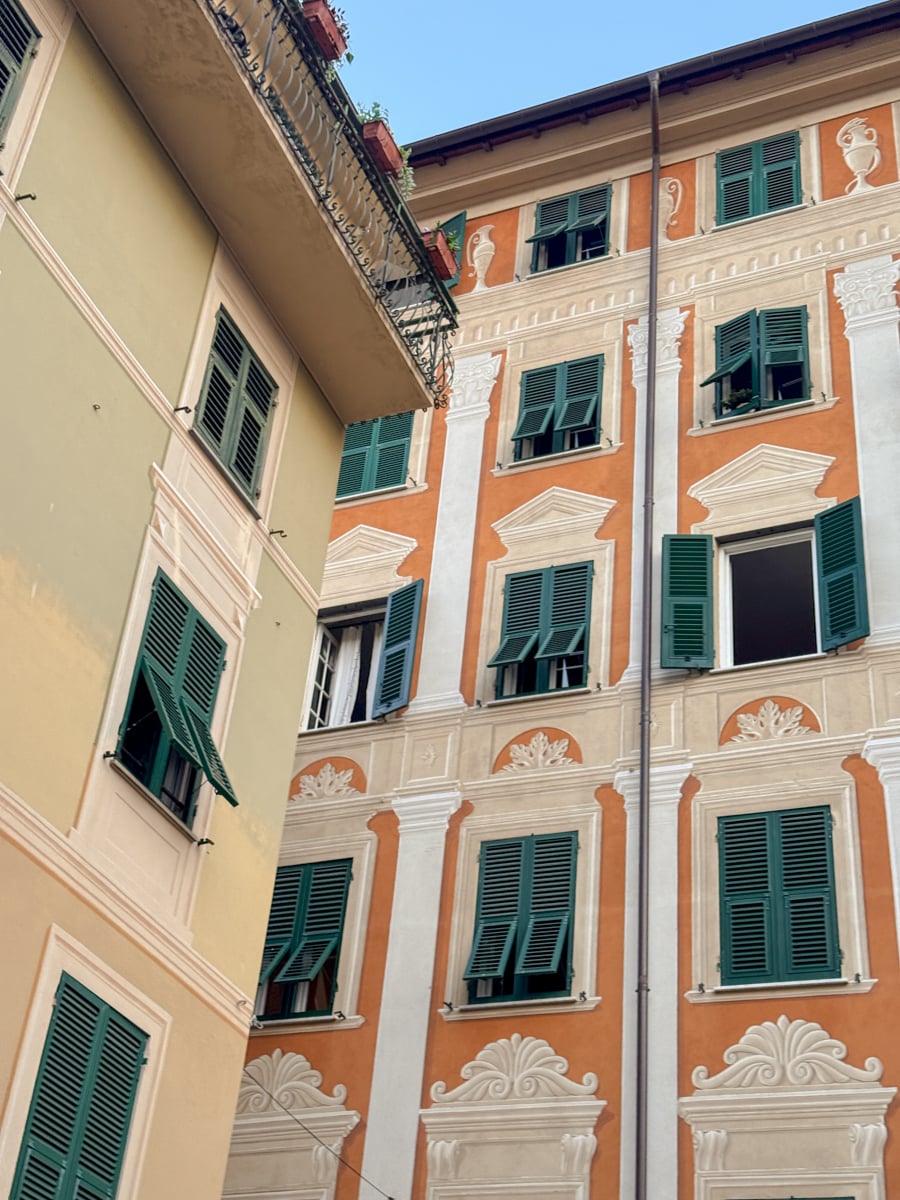 Two colorful buildings with green shuttered windows, one beige and one orange with painted decorative details, are pictured under a clear blue sky in Santa Margherita Ligure. Most windows are open, and some have balconies adorned with plants.