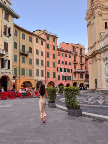 A woman in a beige dress walks through the charming square of Santa Margherita Ligure, surrounded by colorful buildings, outdoor cafes, and ornate architecture under a clear blue sky.