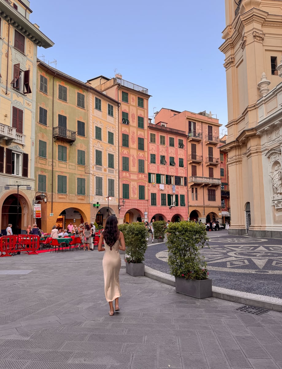 A woman in a beige dress walks through the charming square of Santa Margherita Ligure, surrounded by colorful buildings, outdoor cafes, and ornate architecture under a clear blue sky.