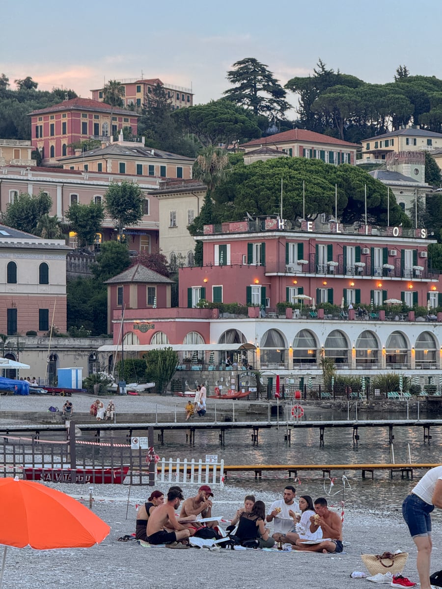 People sit on a pebble beach near an orange umbrella, with a backdrop of colorful buildings, trees, and docks by the waterfront in Santa Margherita Ligure, an enchanting Italian coastal town.