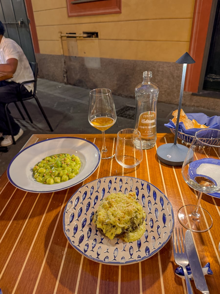 A wooden table set with two plates of food, wine glasses, a clear bottle of water, a bread basket, and a lamp captures the inviting atmosphere of an evening in Santa Margherita Ligure, with another person seated nearby outside on a city street at night.