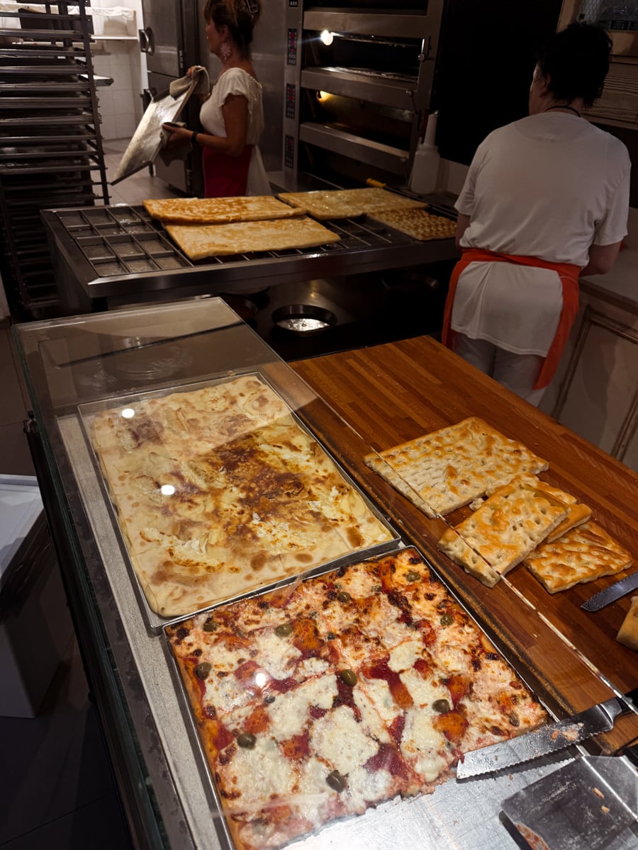 Pizza squares on display behind glass, with two women in white clothing and orange aprons preparing more pizza in the background in a kitchen setting. Various types of pizza are visible on trays and wooden boards, reminiscent of a classic pizzeria in Santa Margherita Ligure.