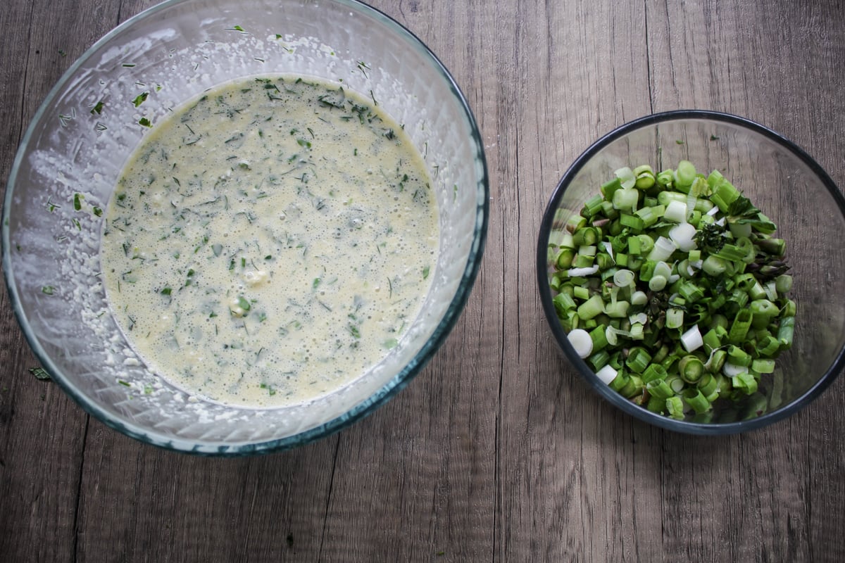 Two glass bowls on a wooden surface: one with a creamy, speckled batter mixed with herbs, and the other with chopped green onions.