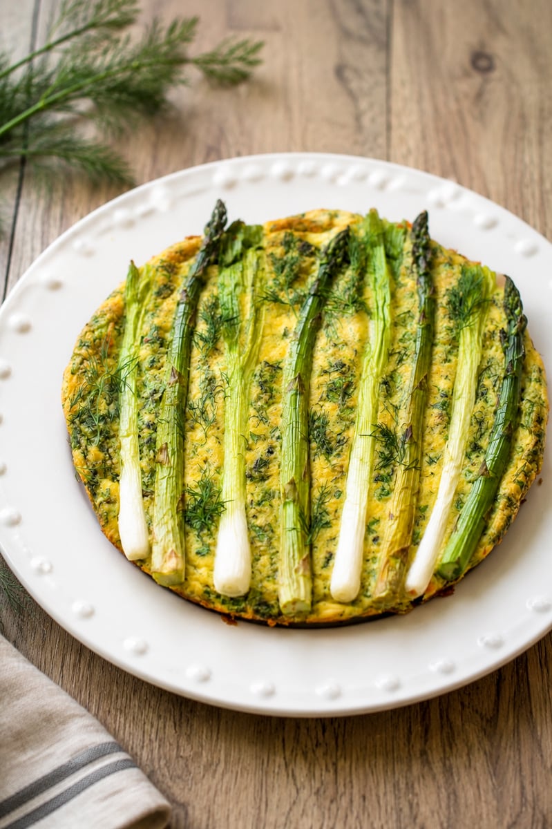 A round frittata topped with whole asparagus spears and dill is served on a white plate with a dotted rim, set on a wooden table with a sprig of dill and a striped cloth nearby.