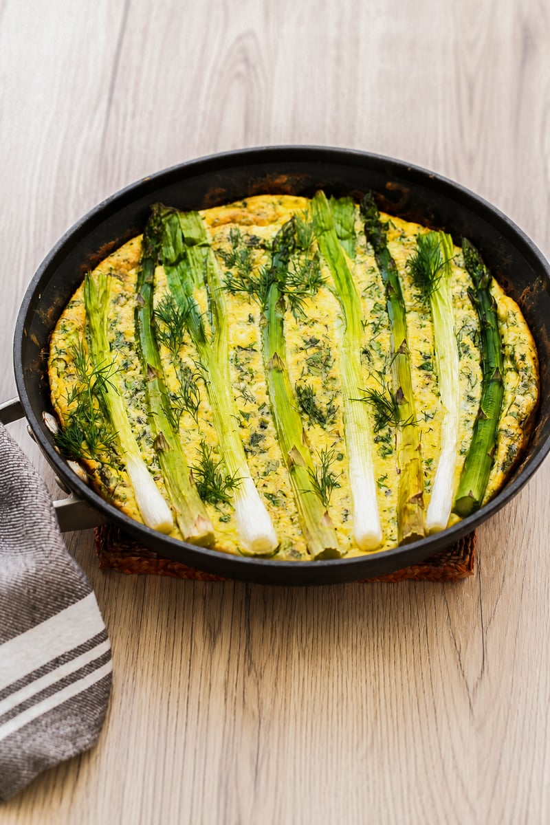 A round pan with a baked frittata topped with whole green onions and fresh dill, set on a wooden surface next to a striped kitchen towel.