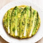 A round frittata topped with whole asparagus spears, served on a white plate with a dotted rim, set on a light wood surface.