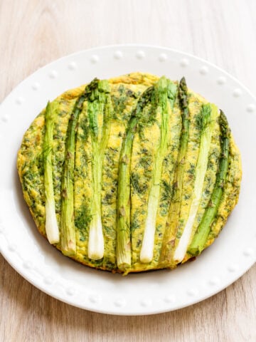 A round frittata topped with whole asparagus spears, served on a white plate with a dotted rim, set on a light wood surface.
