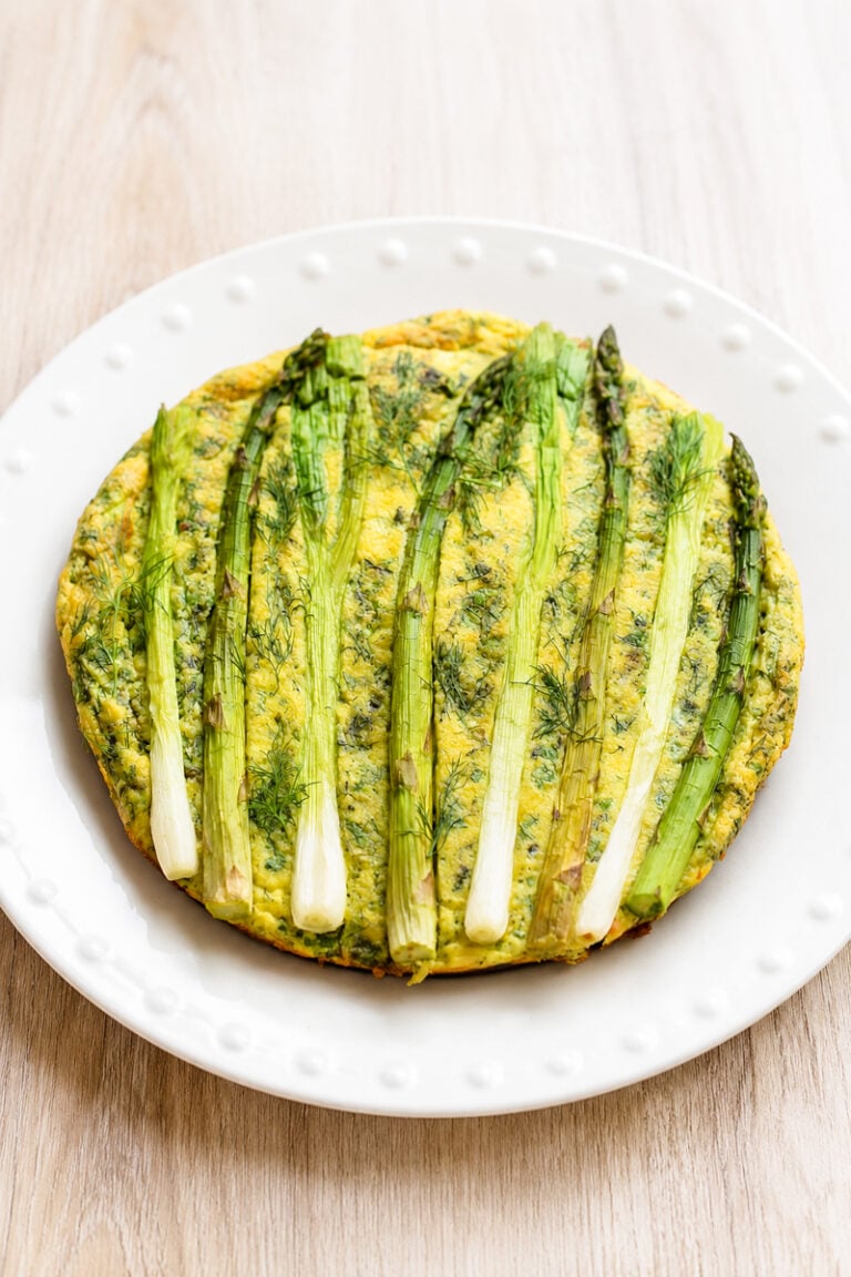 A round frittata topped with whole asparagus spears, served on a white plate with a dotted rim, set on a light wood surface.