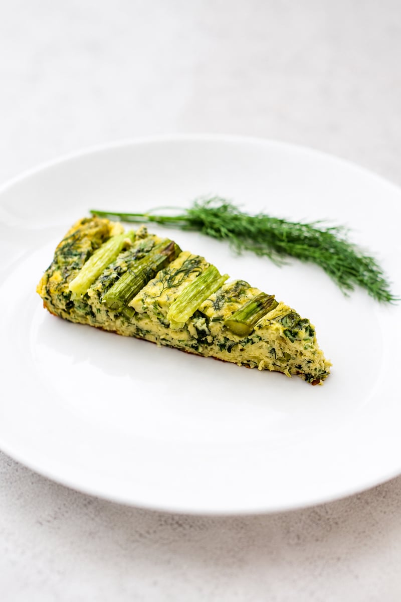 A slice of baked vegetable frittata with visible asparagus, served on a white plate, garnished with fresh dill.