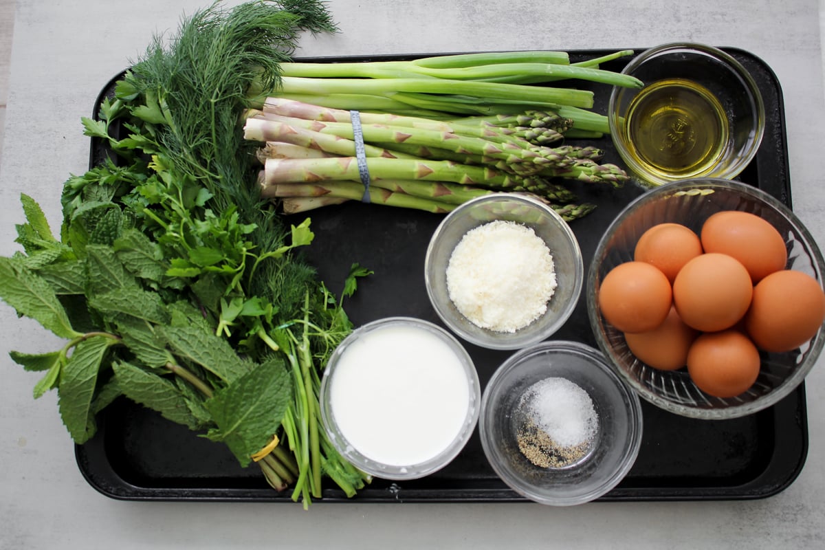 A tray with fresh herbs, green onions, asparagus, a bowl of olive oil, a bowl of eggs, grated cheese, milk, and a small bowl of salt and pepper on a light surface.