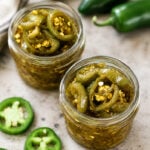 Two glass jars filled with sliced pickled jalapeños in brine, surrounded by fresh whole and sliced jalapeño peppers on a light-colored surface. A cloth napkin is visible in the background.
