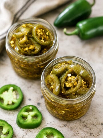 Two glass jars filled with sliced pickled jalapeños in brine, surrounded by fresh whole and sliced jalapeño peppers on a light-colored surface. A cloth napkin is visible in the background.