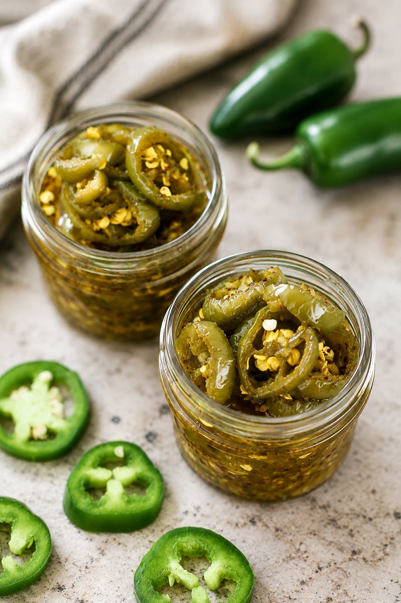 Two glass jars filled with sliced pickled jalape&ntilde;os in brine, surrounded by fresh whole and sliced jalape&ntilde;o peppers on a light-colored surface. A cloth napkin is visible in the background.