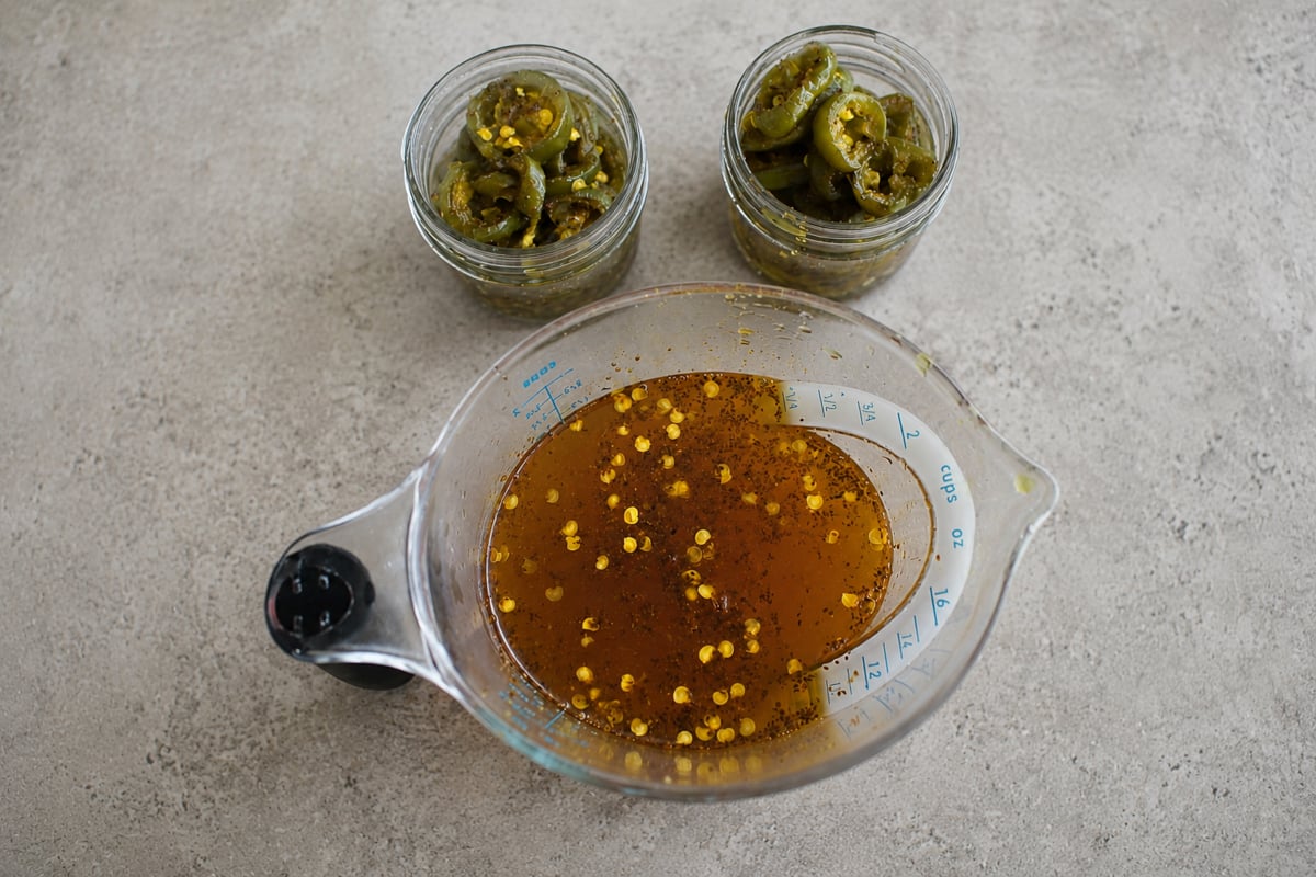 A measuring cup filled with a golden liquid containing pepper seeds sits on a gray surface, next to two small jars filled with sliced pickled jalapeños.