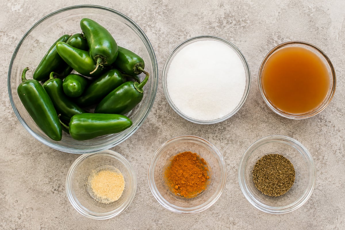 Six glass bowls containing jalapeños, granulated sugar, apple cider vinegar, ground black pepper, turmeric, and garlic powder are arranged on a light countertop.