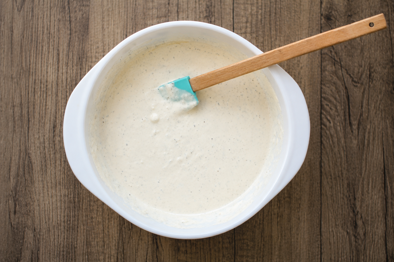 A white mixing bowl filled with creamy pancake batter sits on a wooden surface. A blue spatula with a wooden handle rests inside the bowl.
