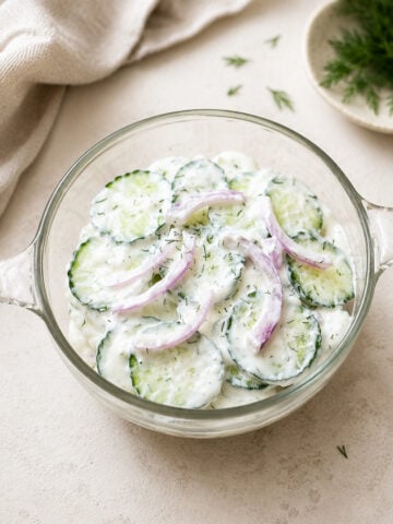 A glass bowl filled with creamy cucumber salad, featuring cucumber slices, thin red onion rings, and fresh dill, sits on a light surface near a beige napkin and a small plate with dill.