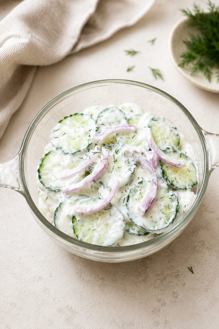 A glass bowl filled with creamy cucumber salad, featuring cucumber slices, thin red onion rings, and fresh dill, sits on a light surface near a beige napkin and a small plate with dill.