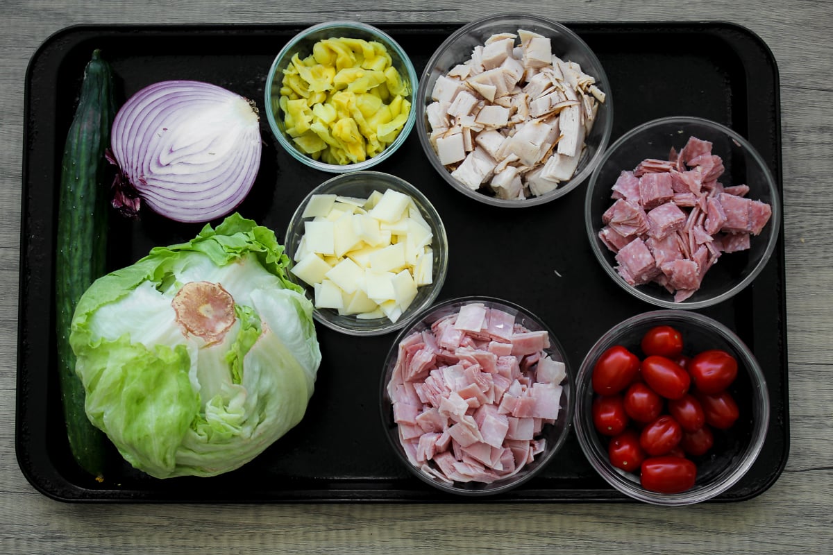 A tray displays a cucumber, half a red onion, a head of iceberg lettuce, cubed cheese, diced yellow peppers, cubed turkey, cubed ham, sliced salami, and grape tomatoes in separate bowls.