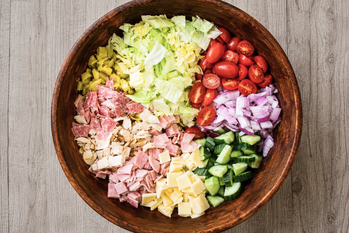 A wooden bowl filled with sections of chopped lettuce, grape tomatoes, red onion, cucumber, cubed cheese, deli meats, and avocado, all neatly separated on a wooden surface.