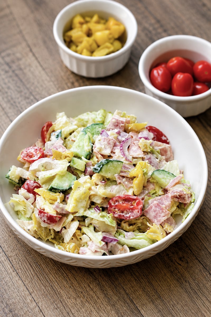 A bowl of chopped salad with lettuce, cucumbers, tomatoes, red onions, and peppers, mixed with dressing. In the background, two small bowls contain sliced yellow peppers and whole cherry tomatoes. The scene is set on a wooden table.