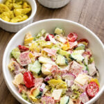 A bowl of chopped salad with lettuce, cucumber, tomato, cheese, salami, and banana peppers on a wooden table, with small bowls of grape tomatoes and banana peppers in the background.
