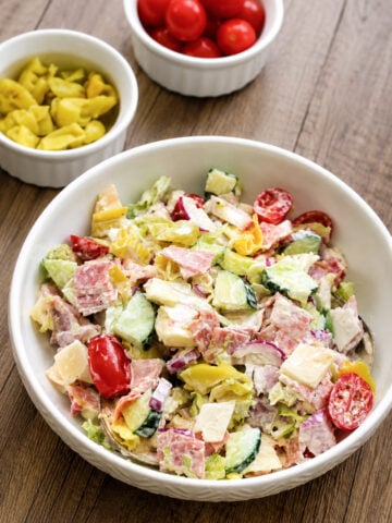 A bowl of chopped salad with lettuce, cucumber, tomato, cheese, salami, and banana peppers on a wooden table, with small bowls of grape tomatoes and banana peppers in the background.