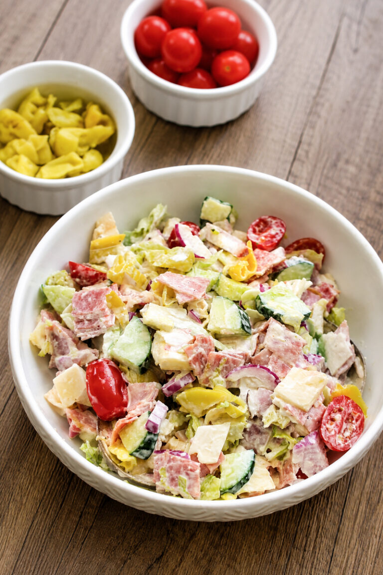 A bowl of chopped salad with lettuce, cucumber, tomato, cheese, salami, and banana peppers on a wooden table, with small bowls of grape tomatoes and banana peppers in the background.