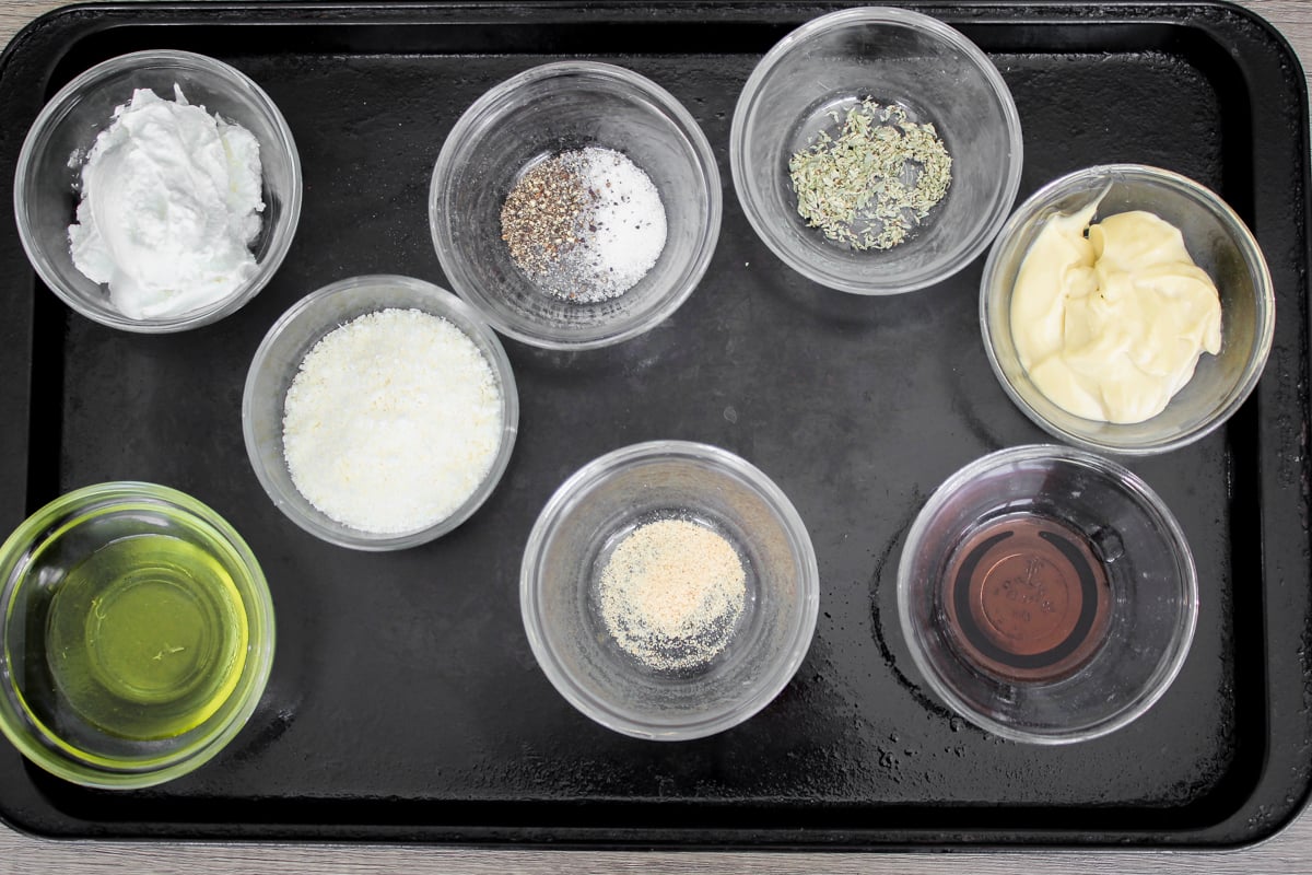 Seven small bowls on a black tray, each containing different ingredients: coconut oil, salt and pepper, dried herbs, mayonnaise, grated parmesan, olive oil, garlic powder, and a dark liquid (possibly vinegar or soy sauce).