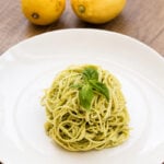 A serving of pesto spaghetti garnished with fresh basil on a white plate, with two yellow lemons in the background on a wooden table.