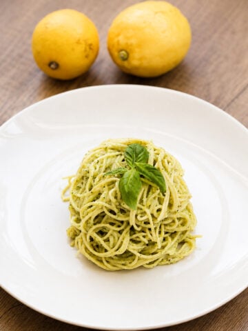 A serving of pesto spaghetti garnished with fresh basil on a white plate, with two yellow lemons in the background on a wooden table.