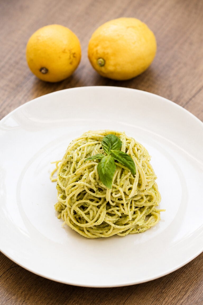 A serving of pesto spaghetti garnished with fresh basil on a white plate, with two yellow lemons in the background on a wooden table.