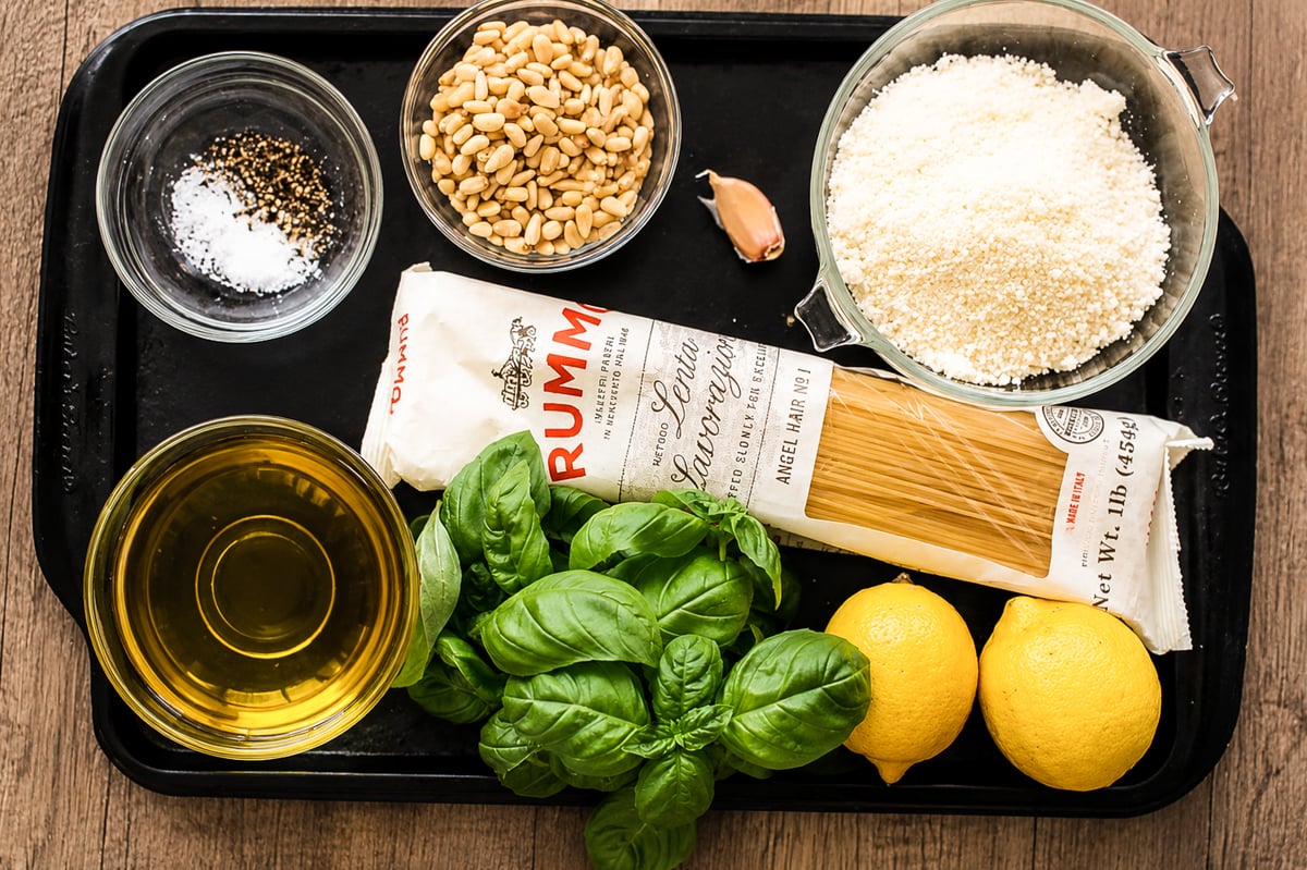 A tray holding olive oil, fresh basil leaves, two lemons, a bundle of dried angel hair pasta, grated parmesan cheese, pine nuts, a clove of garlic, and small bowls of salt and black pepper.