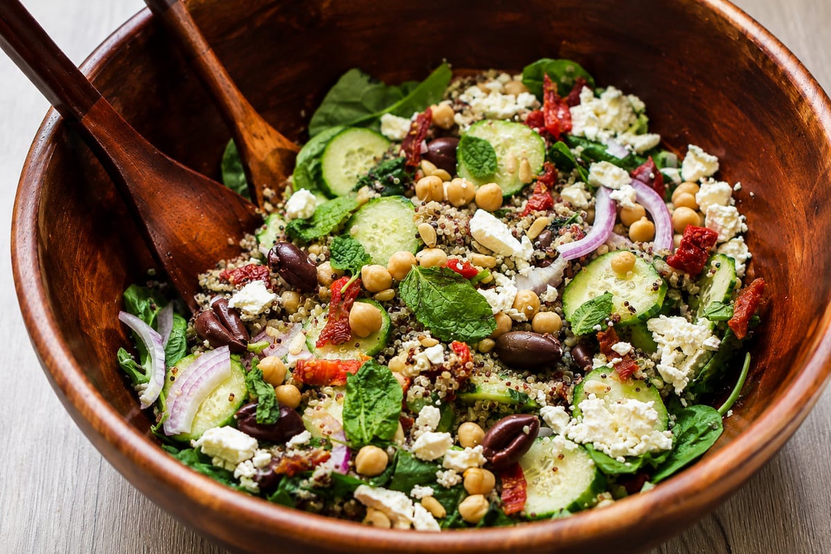A wooden bowl filled with Greek salad featuring cucumbers, chickpeas, red onions, Kalamata olives, feta cheese, sun-dried tomatoes, mint, mixed greens, and quinoa, with wooden salad servers resting inside.