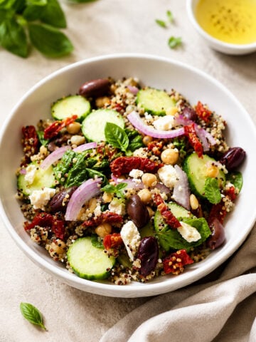 A bowl of Mediterranean quinoa salad with cucumbers, red onions, chickpeas, olives, sun-dried tomatoes, feta cheese, and fresh basil leaves, served with a small dish of dressing on the side.