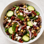 A bowl of Mediterranean quinoa salad with sliced cucumber, sun-dried tomatoes, Kalamata olives, chickpeas, spinach, red onion, and crumbled feta cheese on a light surface.