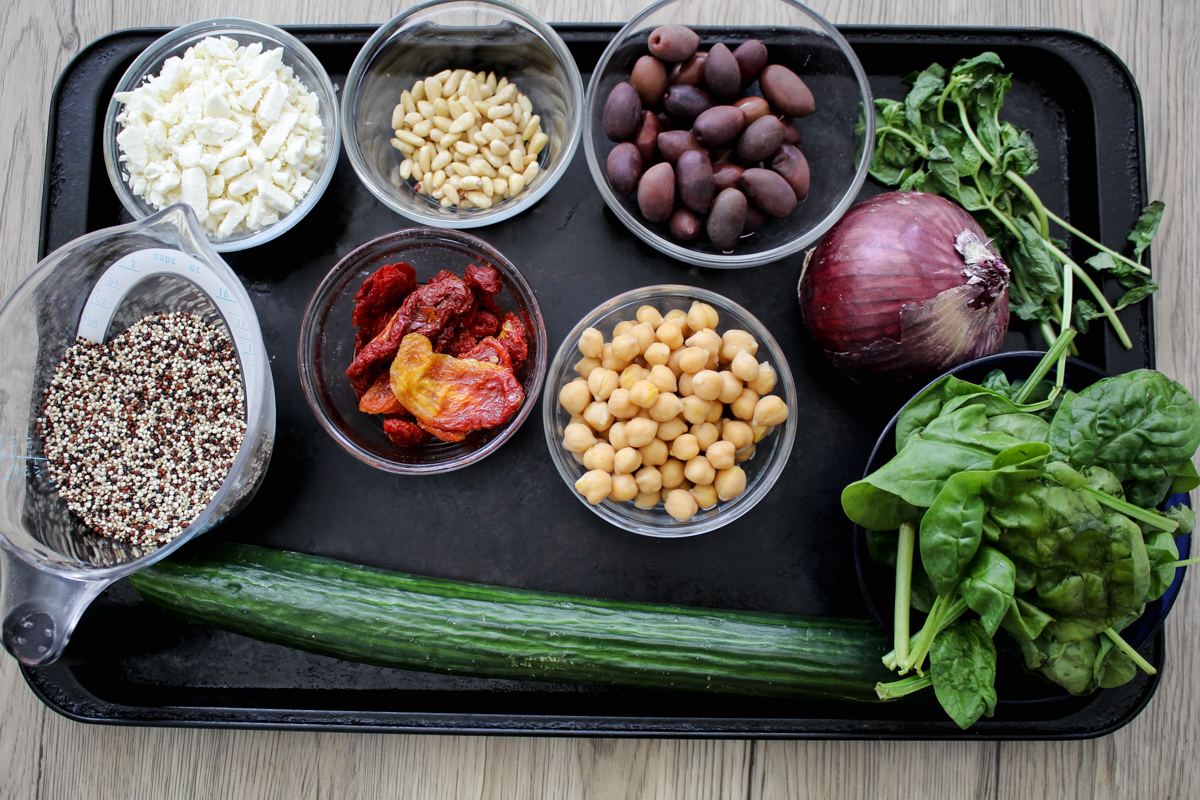 A tray with bowls of quinoa, feta, pine nuts, kalamata olives, sun-dried tomatoes, chickpeas, a red onion, fresh spinach, arugula, and a whole cucumber on a wooden surface.