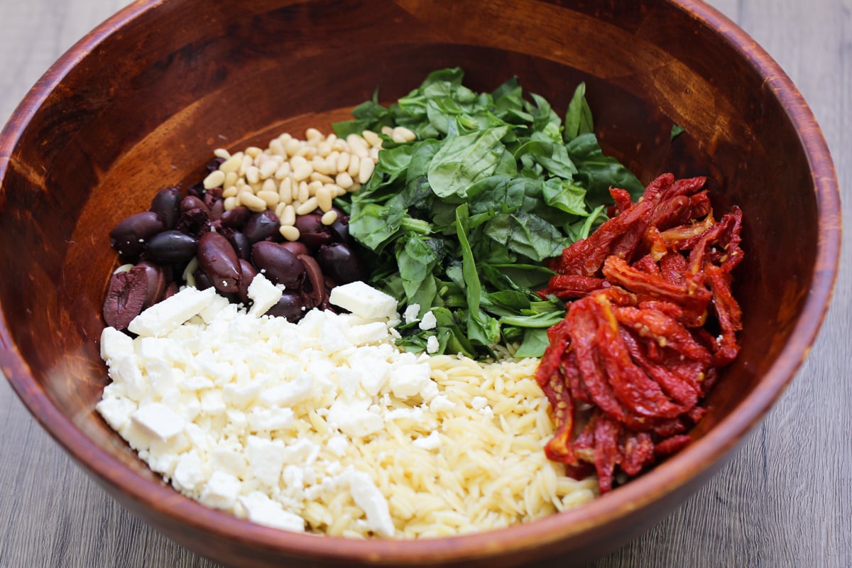 A wooden bowl containing orzo pasta, crumbled feta cheese, sliced Kalamata olives, pine nuts, chopped spinach, and sun-dried tomatoes, arranged in separate sections before mixing.