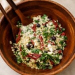 A wooden bowl filled with orzo pasta salad, featuring spinach, sun-dried tomatoes, Kalamata olives, feta cheese, and fresh herbs. Two wooden serving utensils rest inside the bowl.