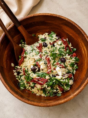A wooden bowl filled with orzo pasta salad, featuring spinach, sun-dried tomatoes, Kalamata olives, feta cheese, and fresh herbs. Two wooden serving utensils rest inside the bowl.