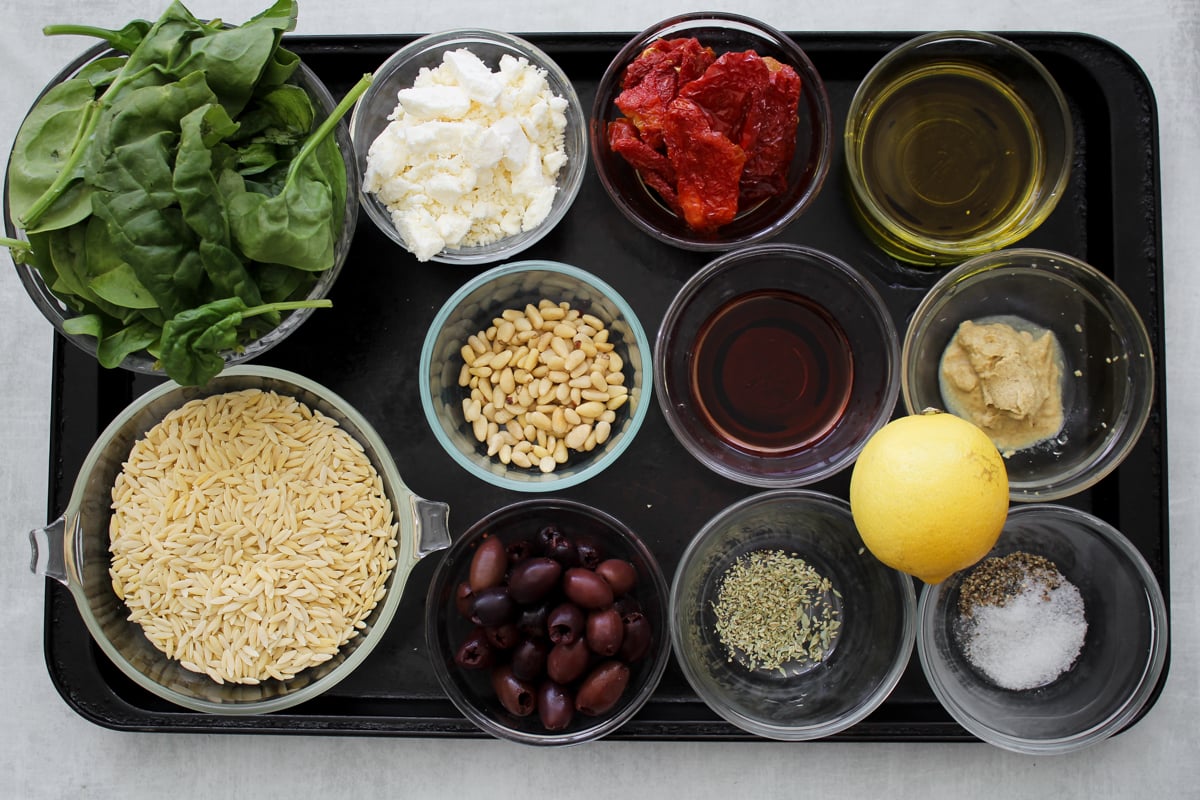 A black tray with bowls of spinach, feta cheese, sun-dried tomatoes, olive oil, orzo pasta, pine nuts, balsamic vinegar, Dijon mustard, Kalamata olives, dried herbs, black pepper, salt, and a lemon.